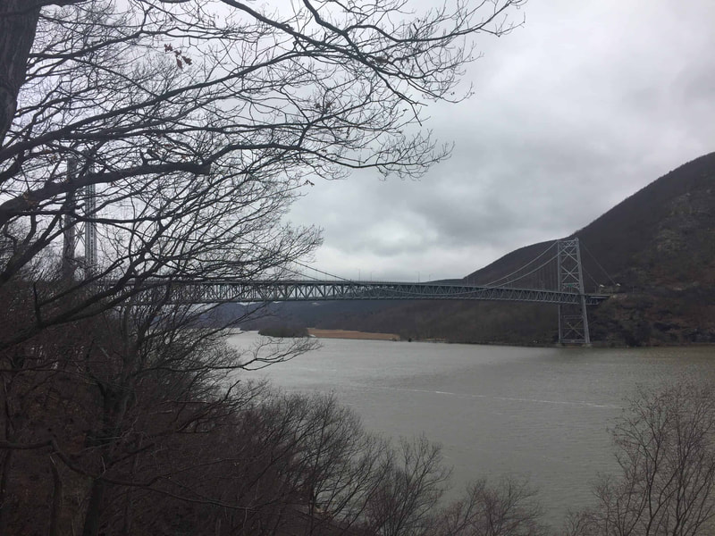 View of river, suspension bridge, mountain and gray sky.