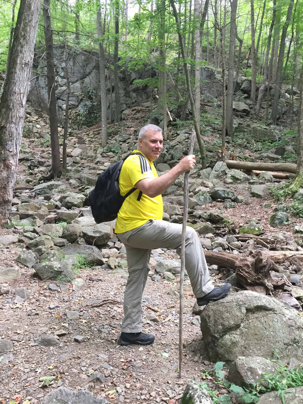 Man in beige pants and yellow shirt with a black backpack carrying a stick and standing alongside a trail.