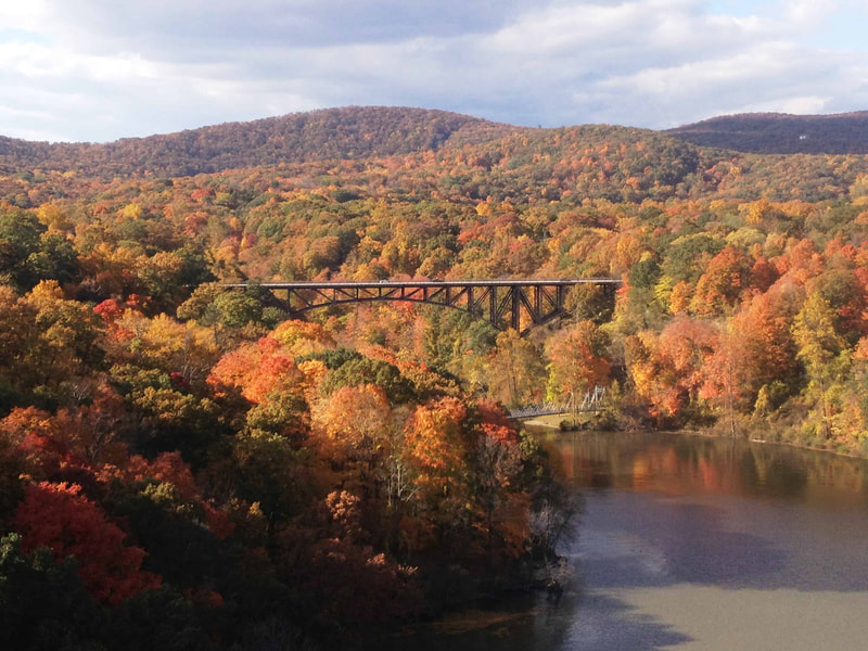Green, red, orange and yellow trees beneath a cloudy sky surround an elevated railroad bridge and a river.