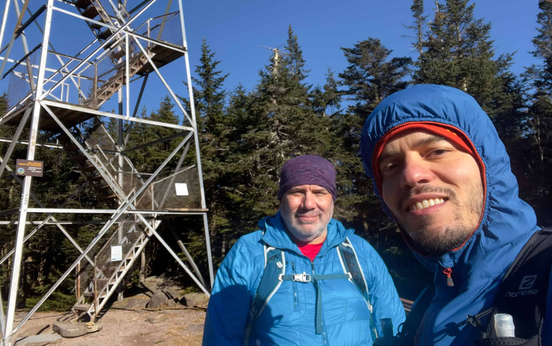 Two men dressed in blue standing on a mountain summit with a steel fire tower, green trees, and blue sky in the background.