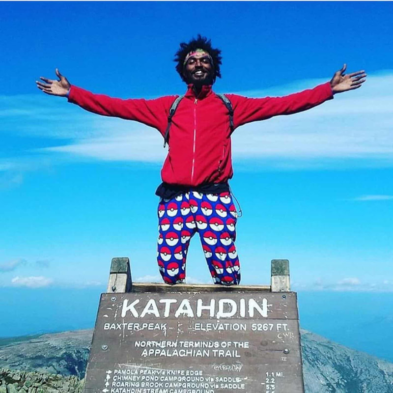 Man in red jacket and colorful pants standing on the Appalachian Trail terminus with arms outstretched in front of a blue sky.