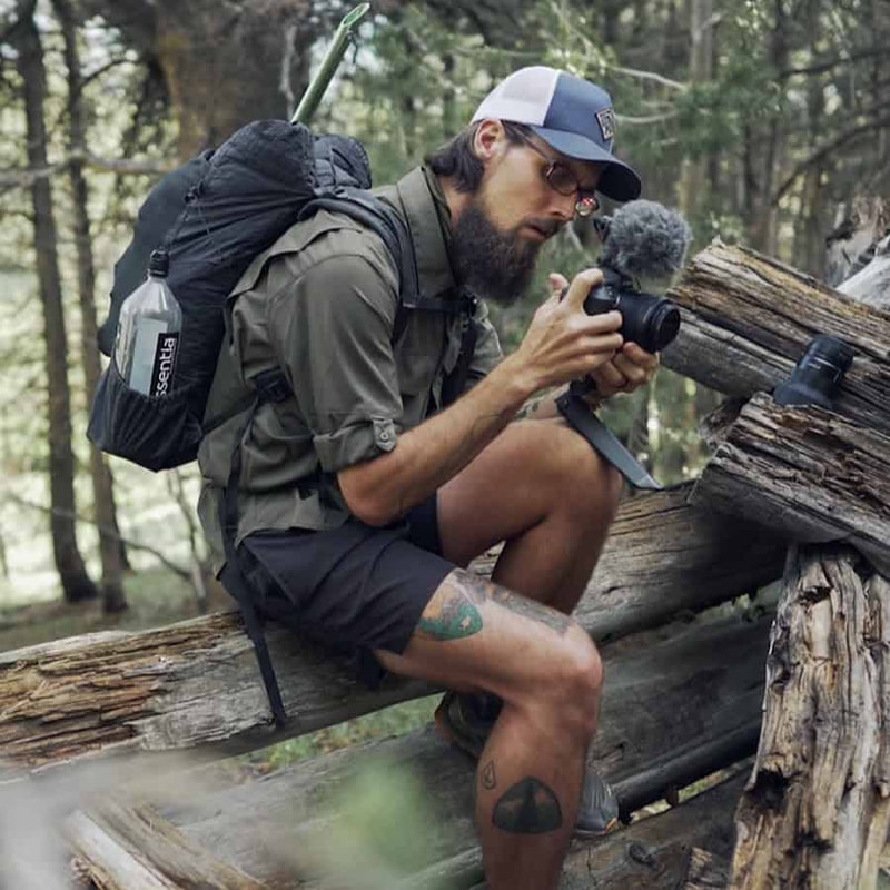 Man with backpack sitting on a fallen tree looking at a camera