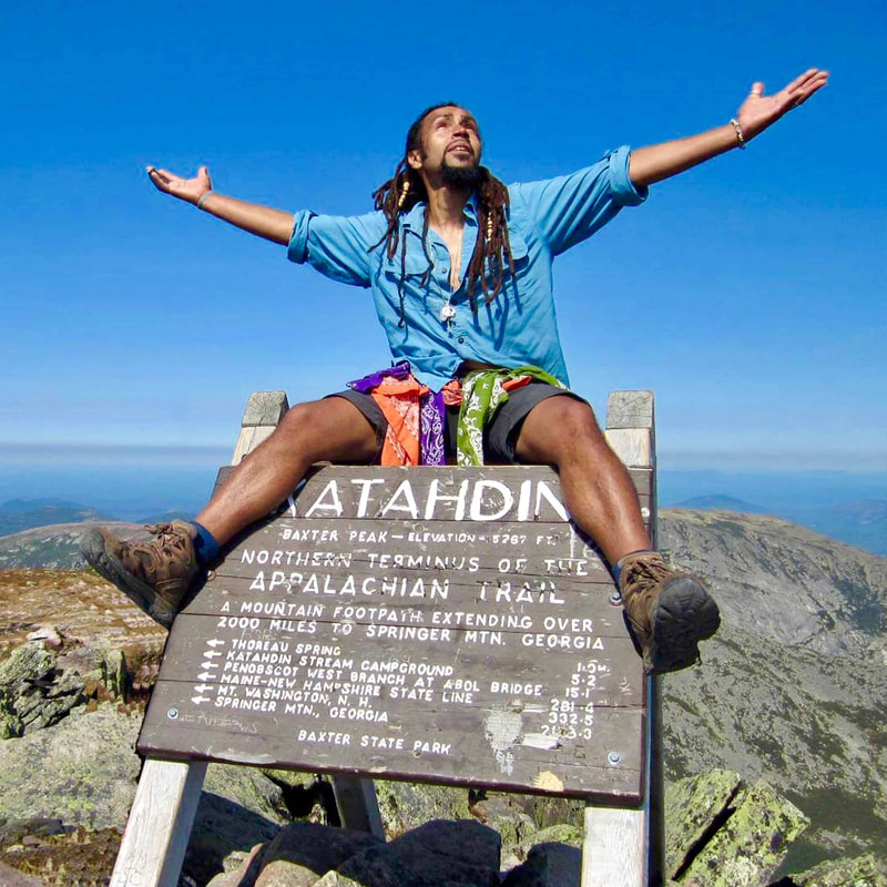 Man in blue shirt, gray shorts, and brown boots sitting on sign atop a mountain with arms outstretched.