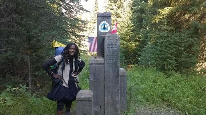 Woman dressed in white and black with backpack standing next to a stone monument marking the terminus of the Continental Divide Trail.