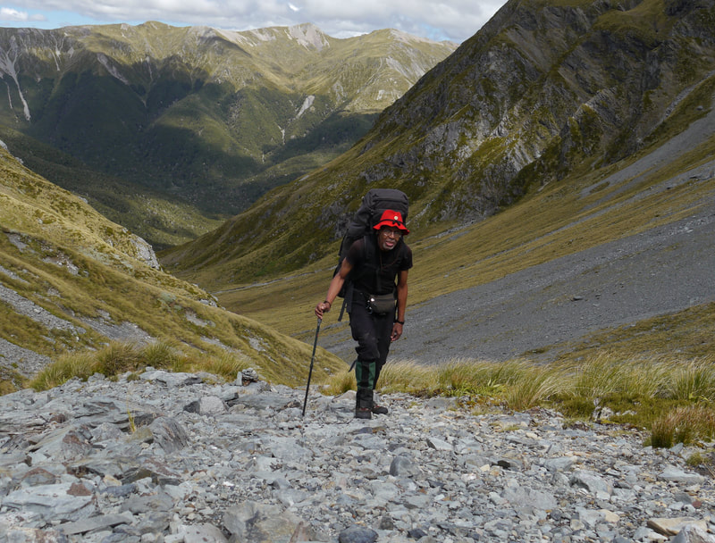 Man in black clothing and red hat with trekking pole and mountains in the background.