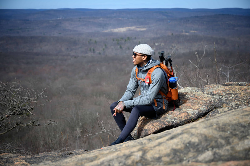 Man with gray jacket and hat, sunglasses, and orange backpack sitting on a mountain with blue sky in the background.