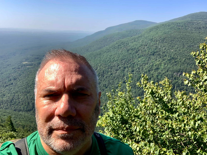 Man with green shirt standing on a mountain summit with green hills and hazy blue sky in the background.